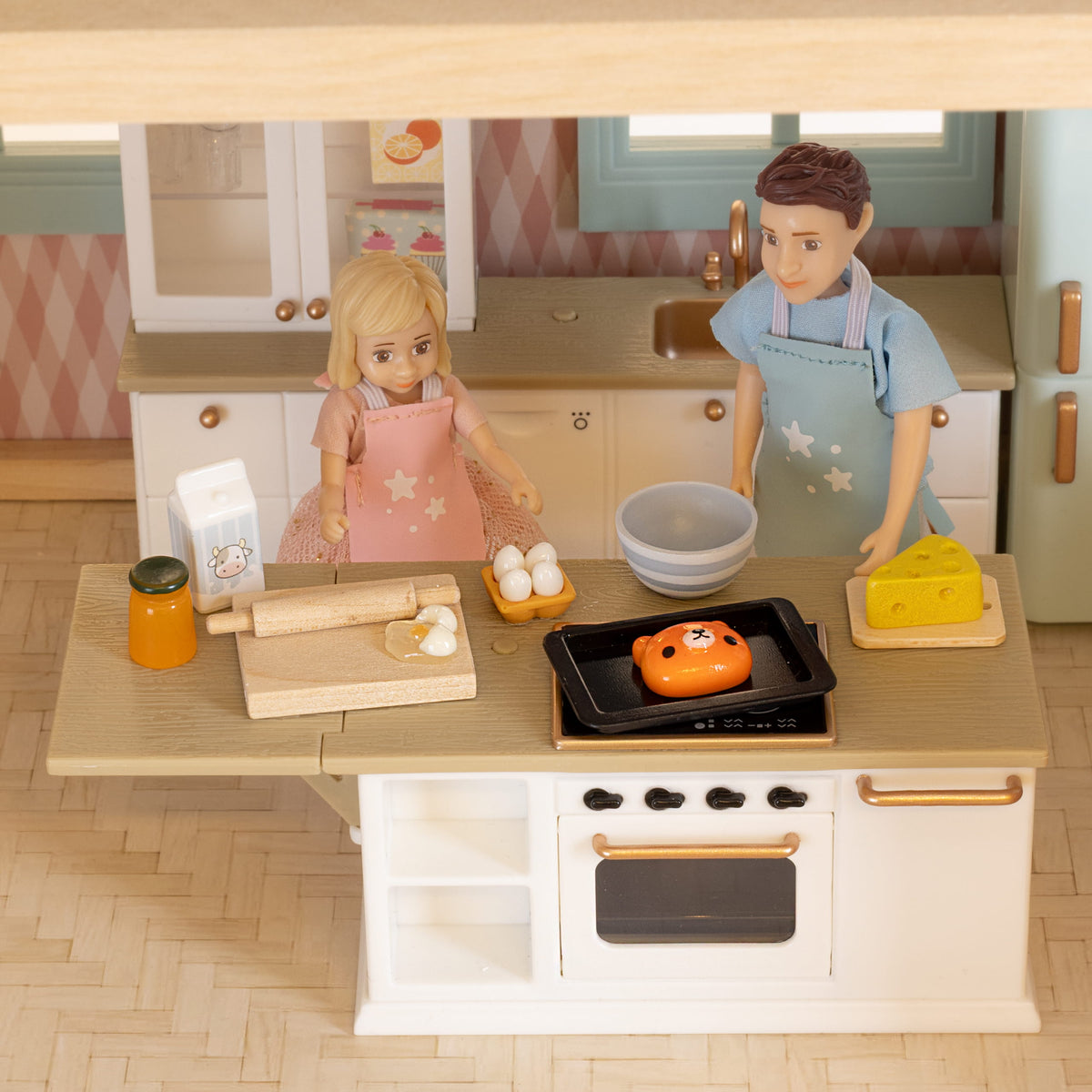 Lundby dolls house dolls in aprons making breakfast in the kitchen, with eggs, cheese, a mixing bowl and a baking tray on the stove as part of their morning routine.